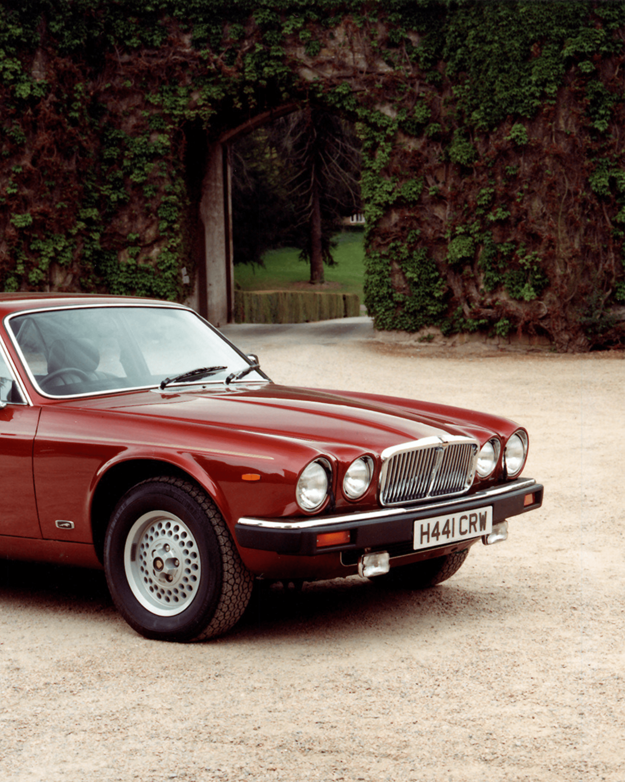Classic red Jagiar parked on gravel path in front of ivy-covered archway.