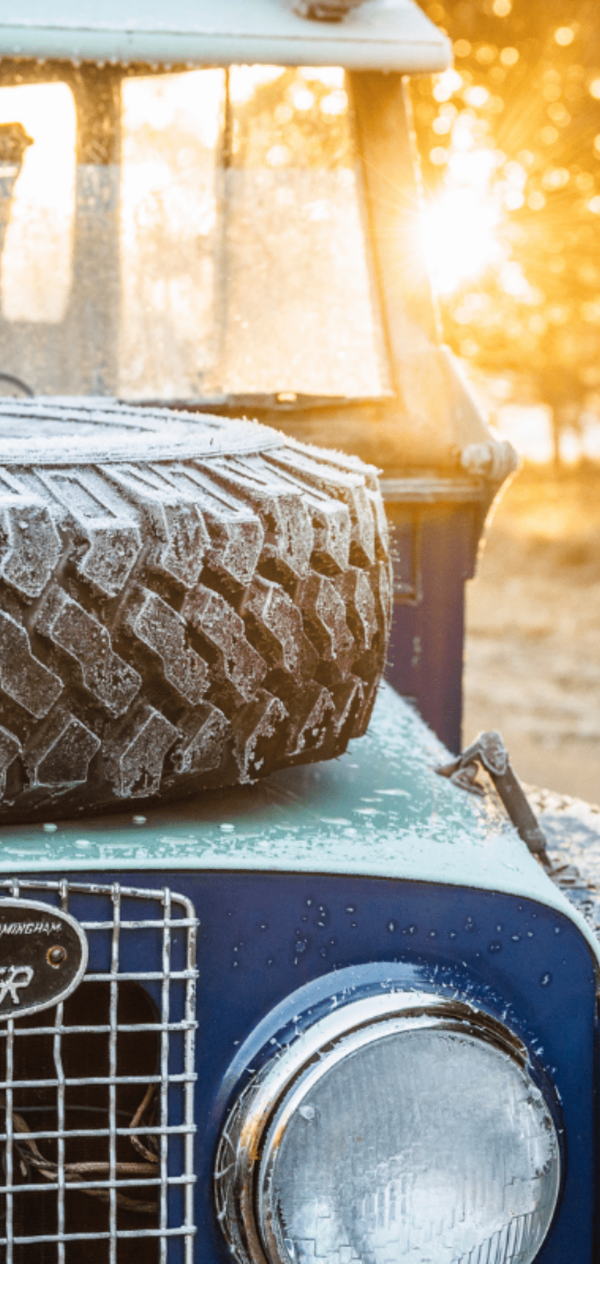 Frost-covered vintage Land Rover with hood-mounted spare tire in forest setting.