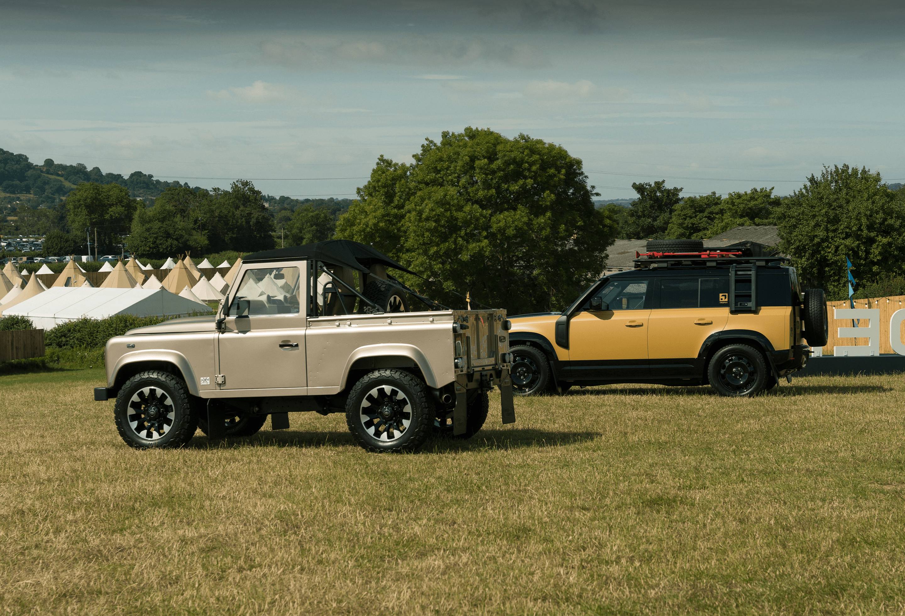 Two off-road Defenders parked on a grassy field with tents and trees in the background.