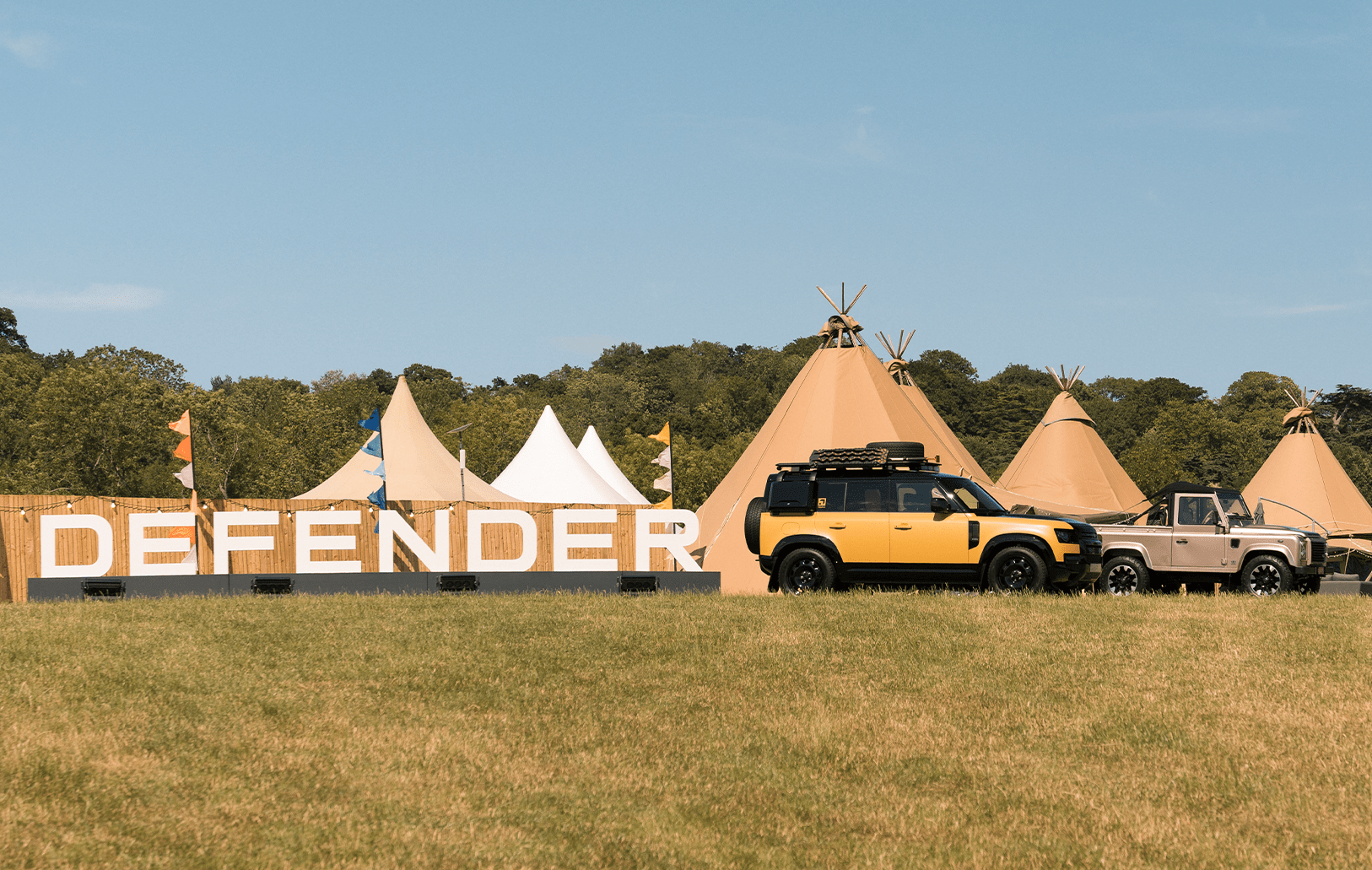 DEFENDER sign with two vehicles in front of teepee tents on a grassy field under a clear sky.