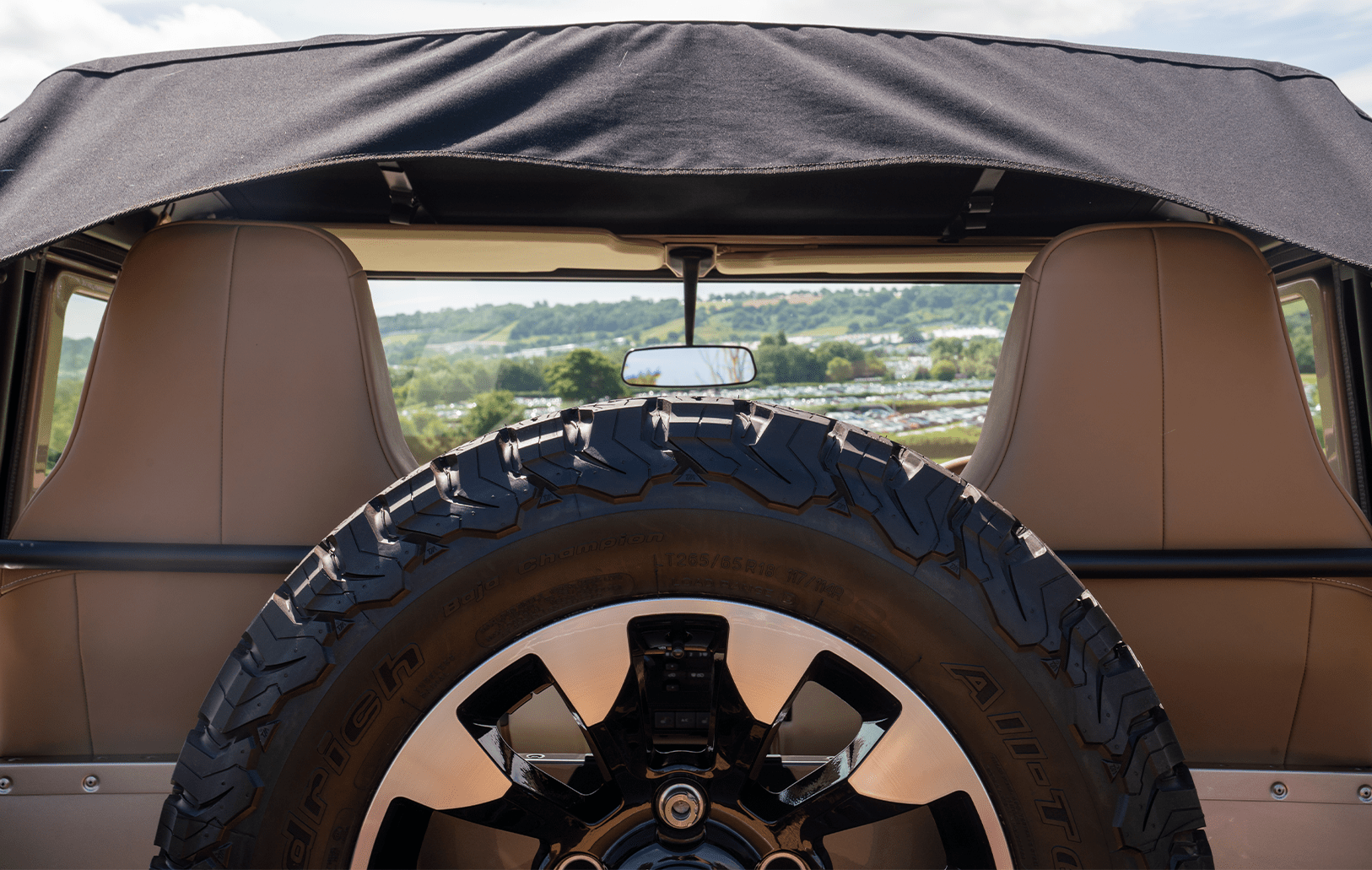 Rear view of a classic Defedner with spare tire and soft top, set against green hills and a cloudy sky.