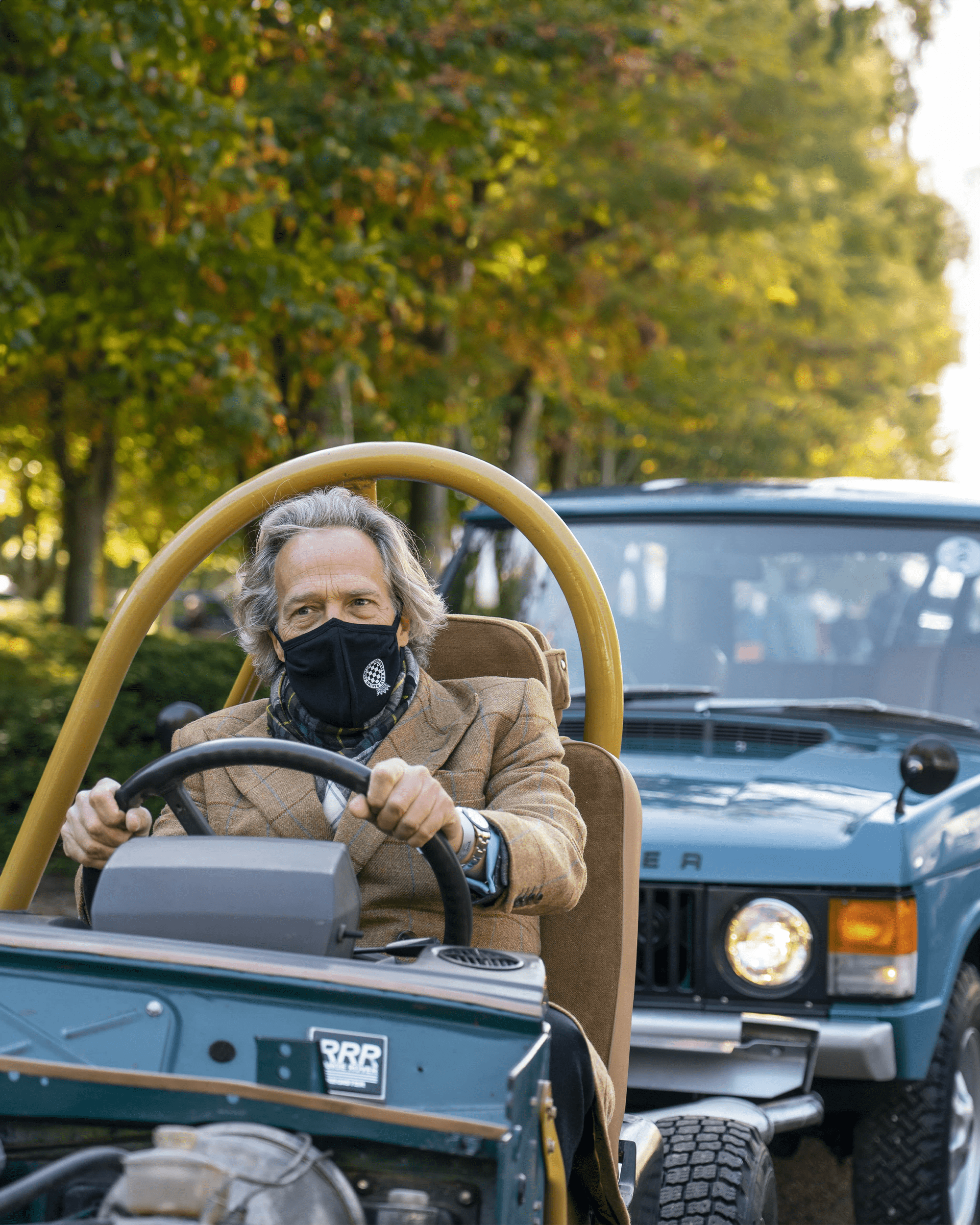 Off-road vehicle with roll cage driving through wooded area, classic Range Rover behind.