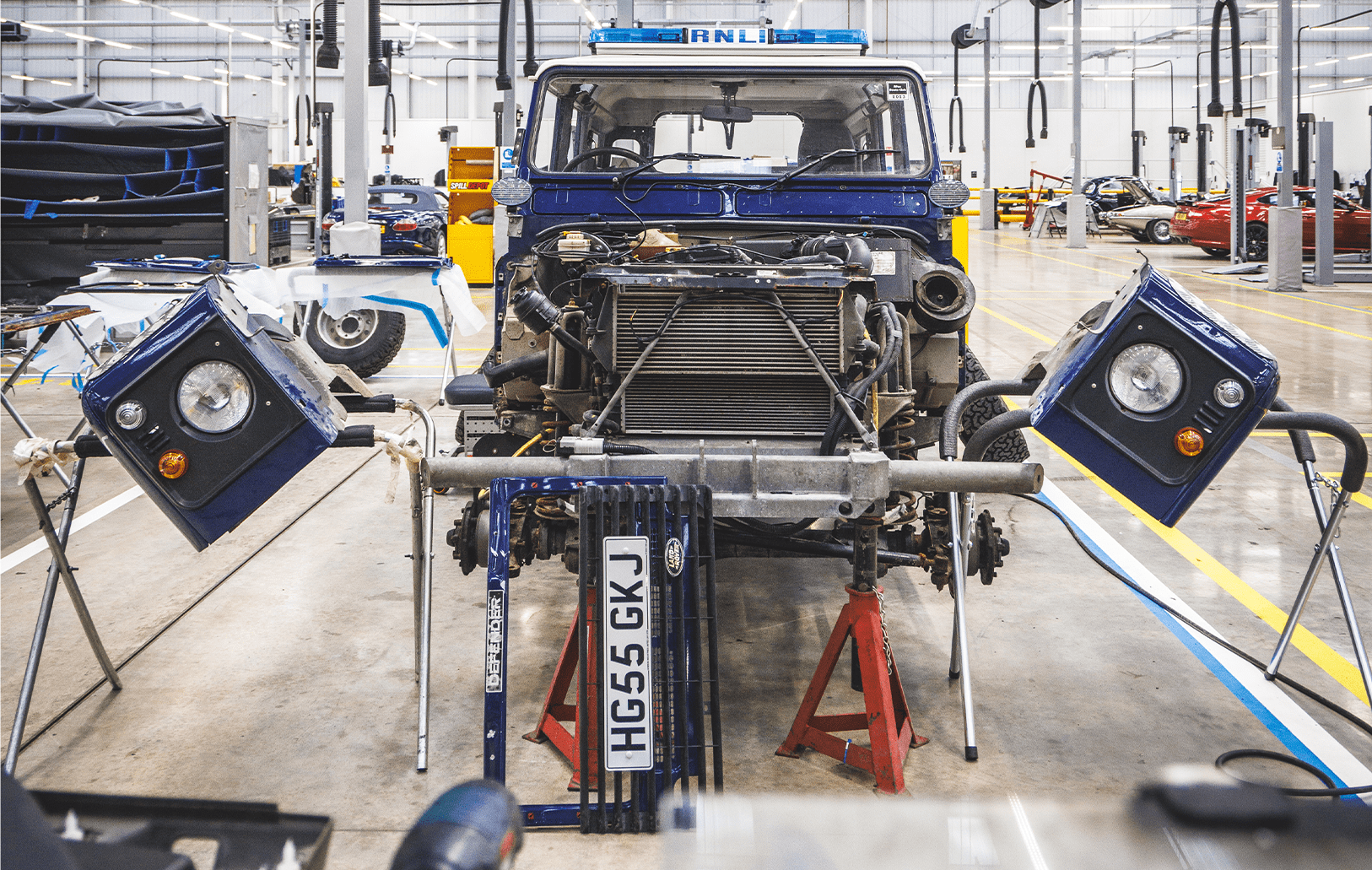 Partially disassembled blue vehicle on jack stands in workshop, with exposed engine and headlight assemblies.