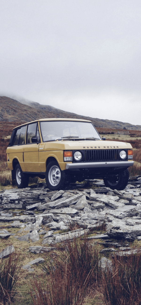 Yellow Range Rover on rocky terrain beside stone structure, with hills in background.