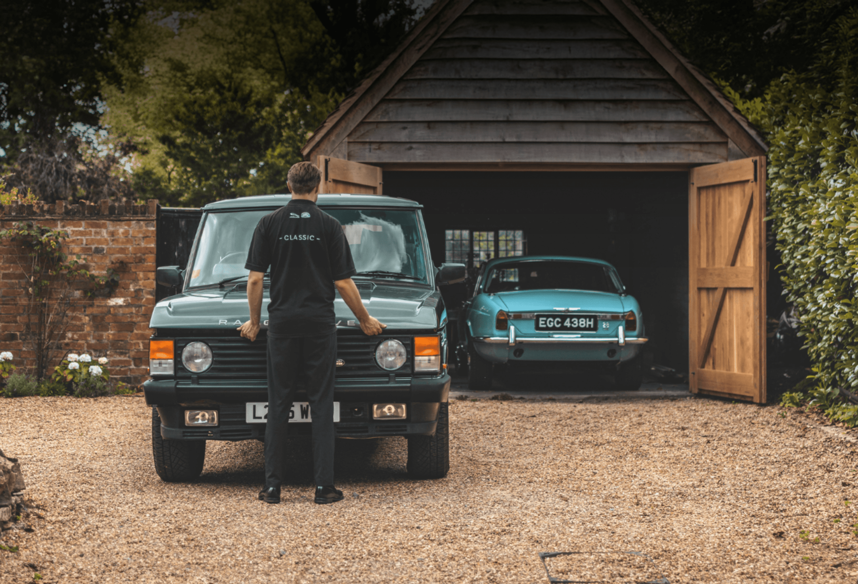 Mechanic in “CLASSIC” uniform standing by green Range Rover near open garage with blue classic Jaguar inside.