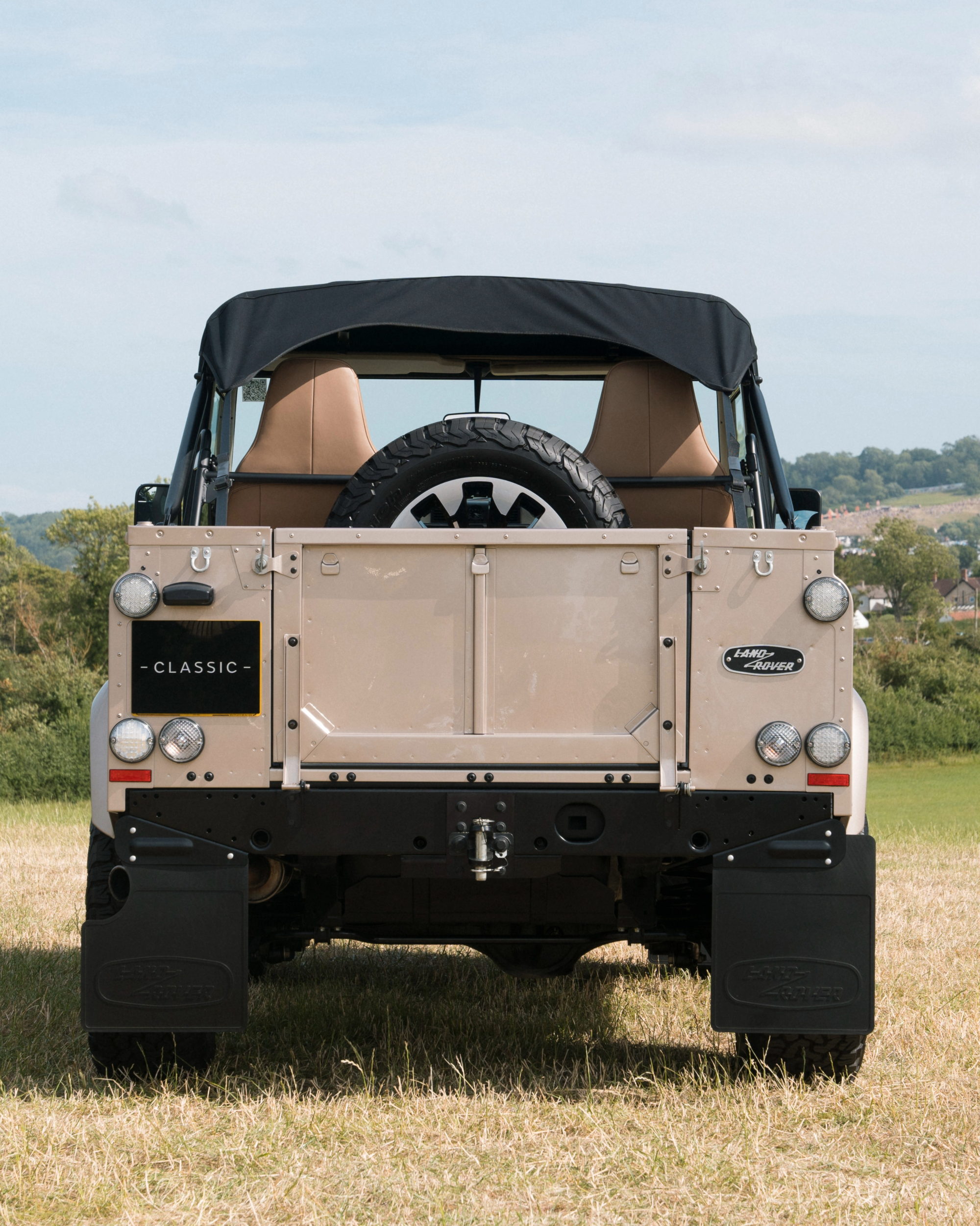 Rear view of a beige Land Rover Classic with soft top and spare tire, parked on grass.