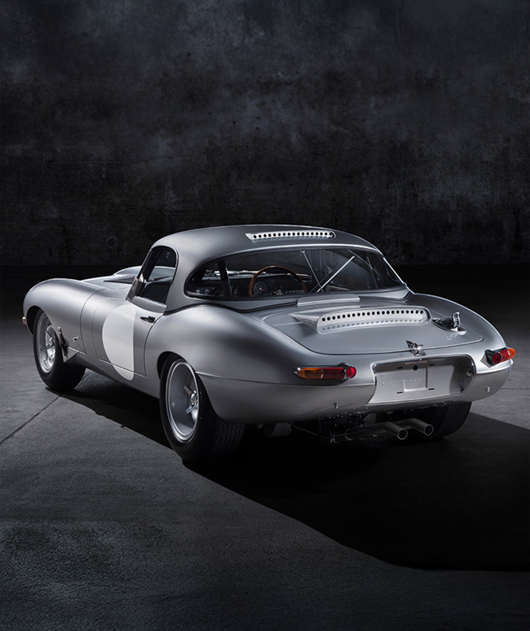 Classic silver E-Type, rear three-quarter view against dark background.