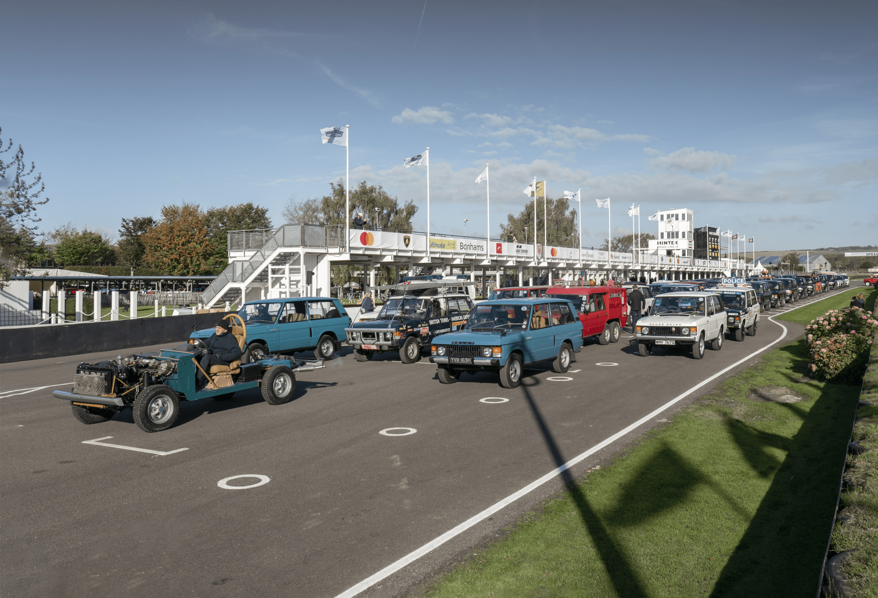 Lineup of vintage Land Rovers on racetrack with flags and viewing structure.