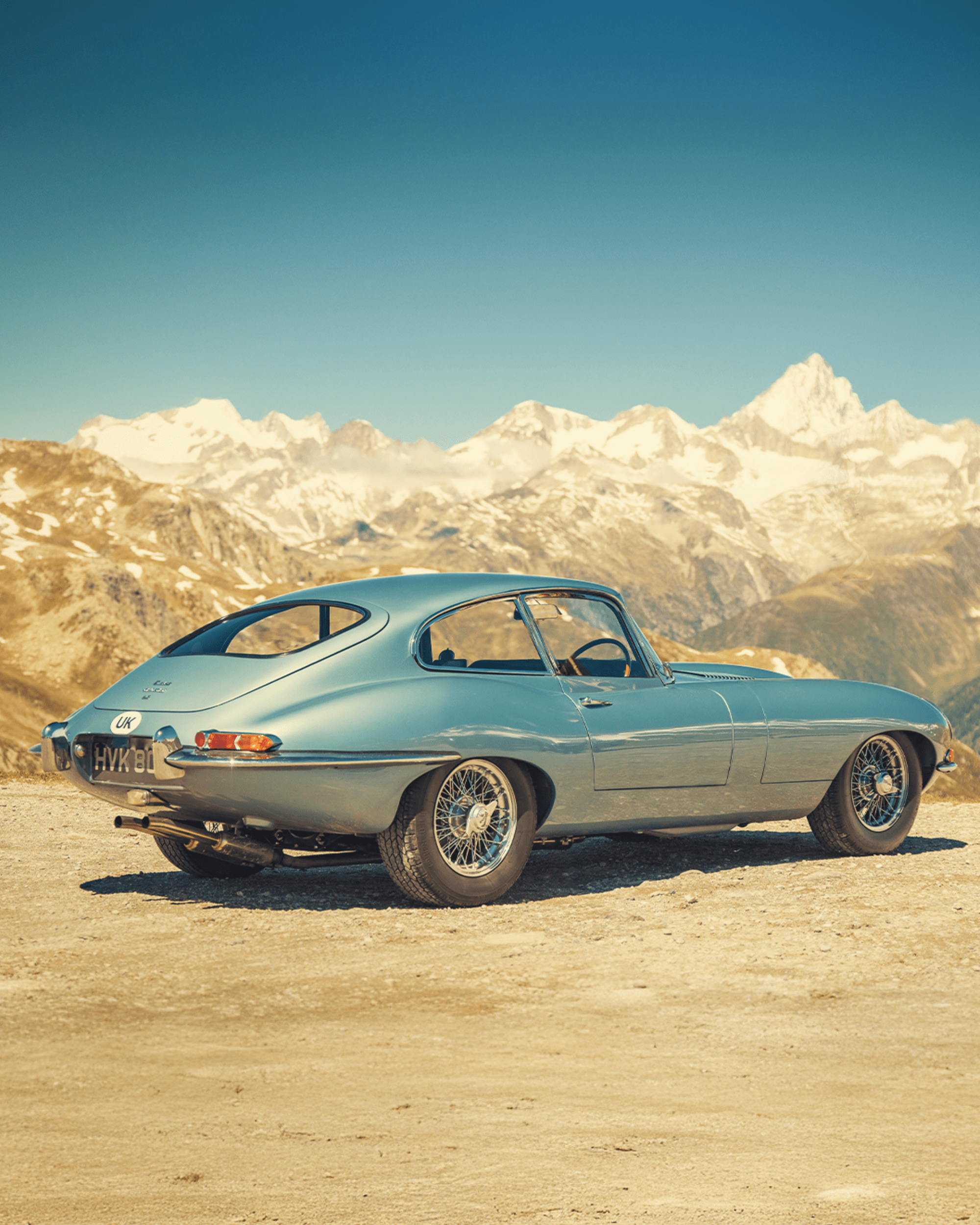 Classic blue Jaguar on dirt road with snow-covered mountains in the background.